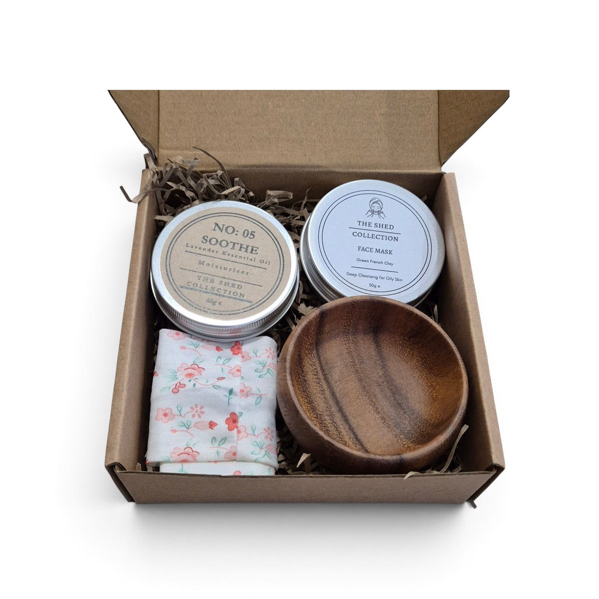 A gift set in an open brown cardboard box with shredded paper filler, containing two round metal tins, a folded floral headband and a small wooden bowl.