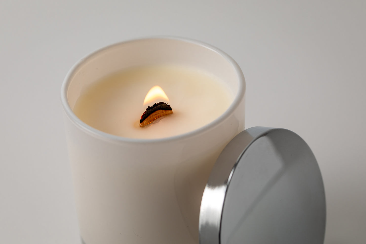 Picture shows vegan friendly candle from The Shed Collection in a white jar. The wooden wick is burning with a low flame.