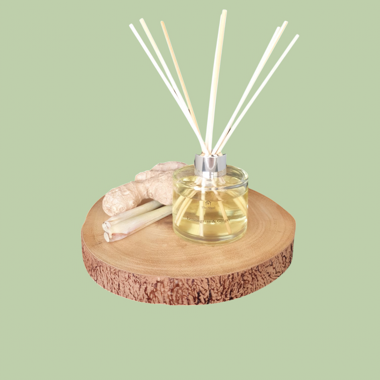 Picture shows a vegan friendly Lemongrass & Ginger Reed Diffuser from The Shed Collection on a log slice beside pieces of fresh lemongrass and ginger.