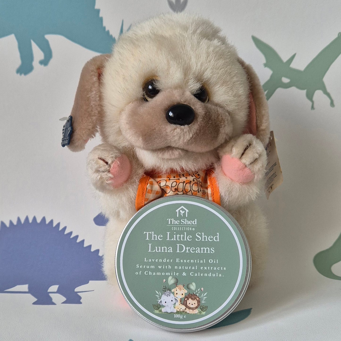 A plush cream-colored puppy toy holding a round tin of 'The Shed Collection' 'The Little Shed Luna Dreams' serum. The background is white with dinosaur silhouettes in muted blue and green.