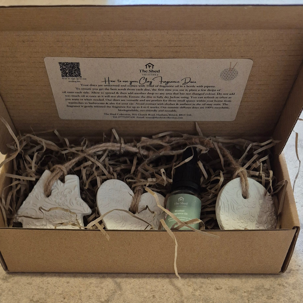 An open brown cardboard gift box filled with shredded paper and containing a small green bottle and three white ceramic ornaments, with a printed instruction card inside the lid.