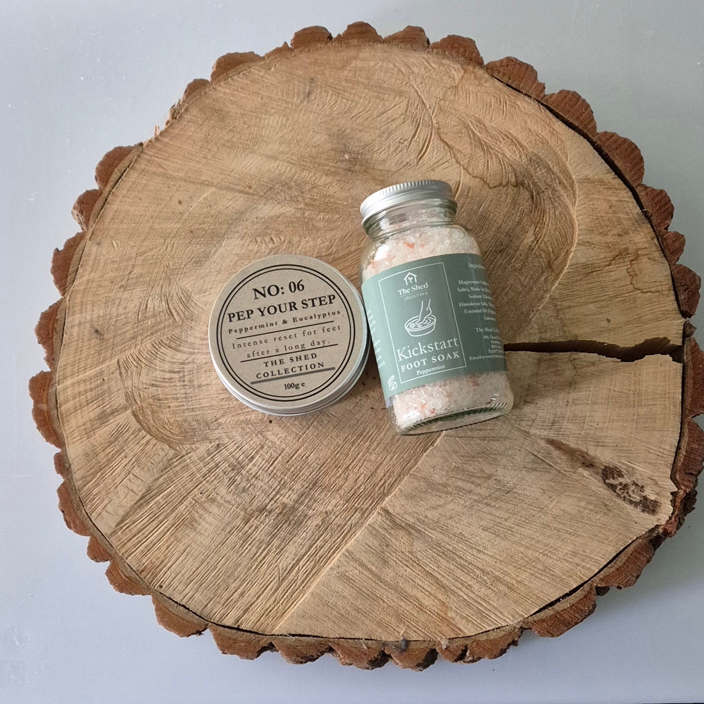 A jar of Peppermint Kickstart Bath Salts and a round container of Pep Your Step Foot Cream on a log slice with a light background.
