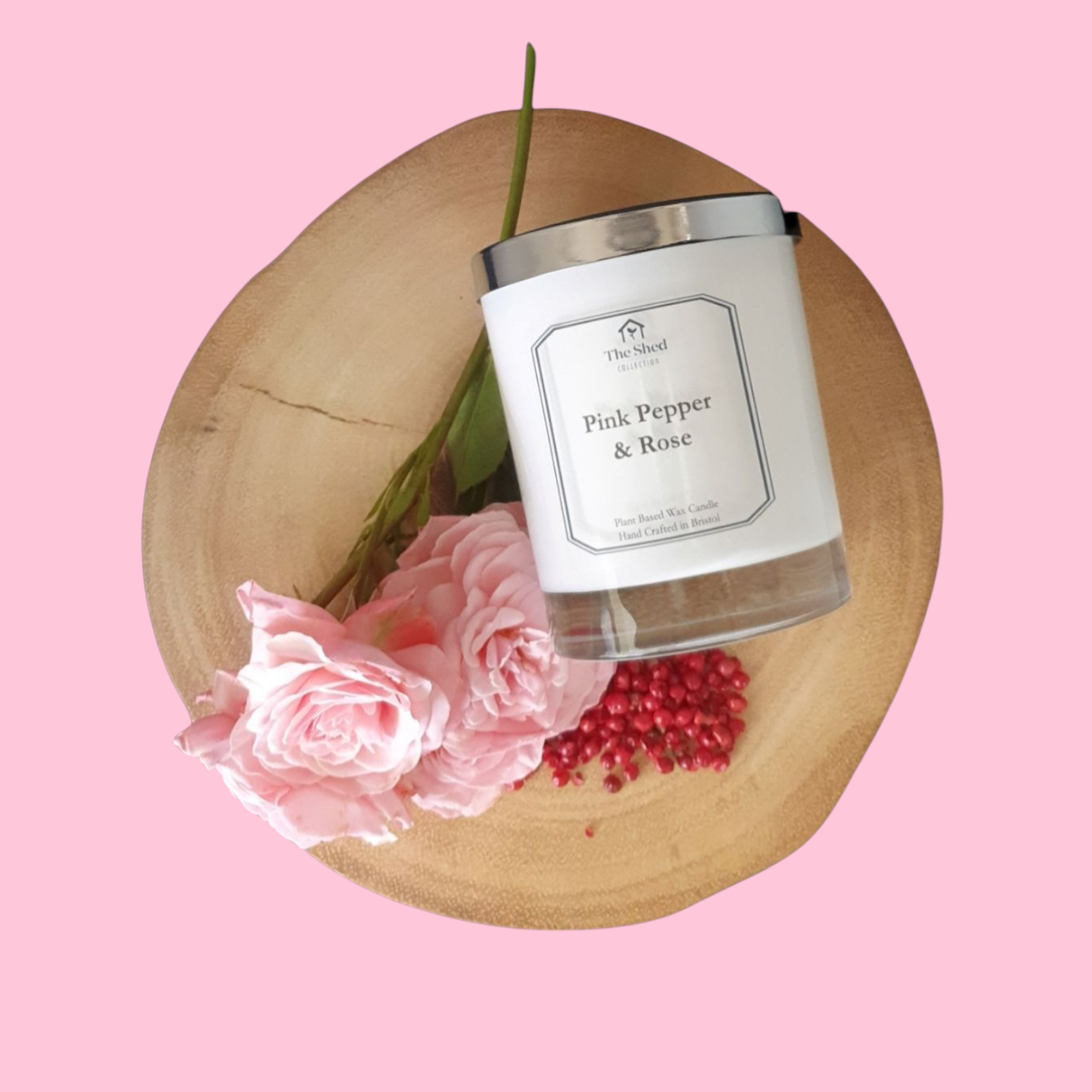 Picture shows a vegan friendly Pink Pepper & Rose candle from The Shed Collection in a white container with silver lid on a log slice beside freshly cut pink tea roses and some vibrant pink peppercorns.