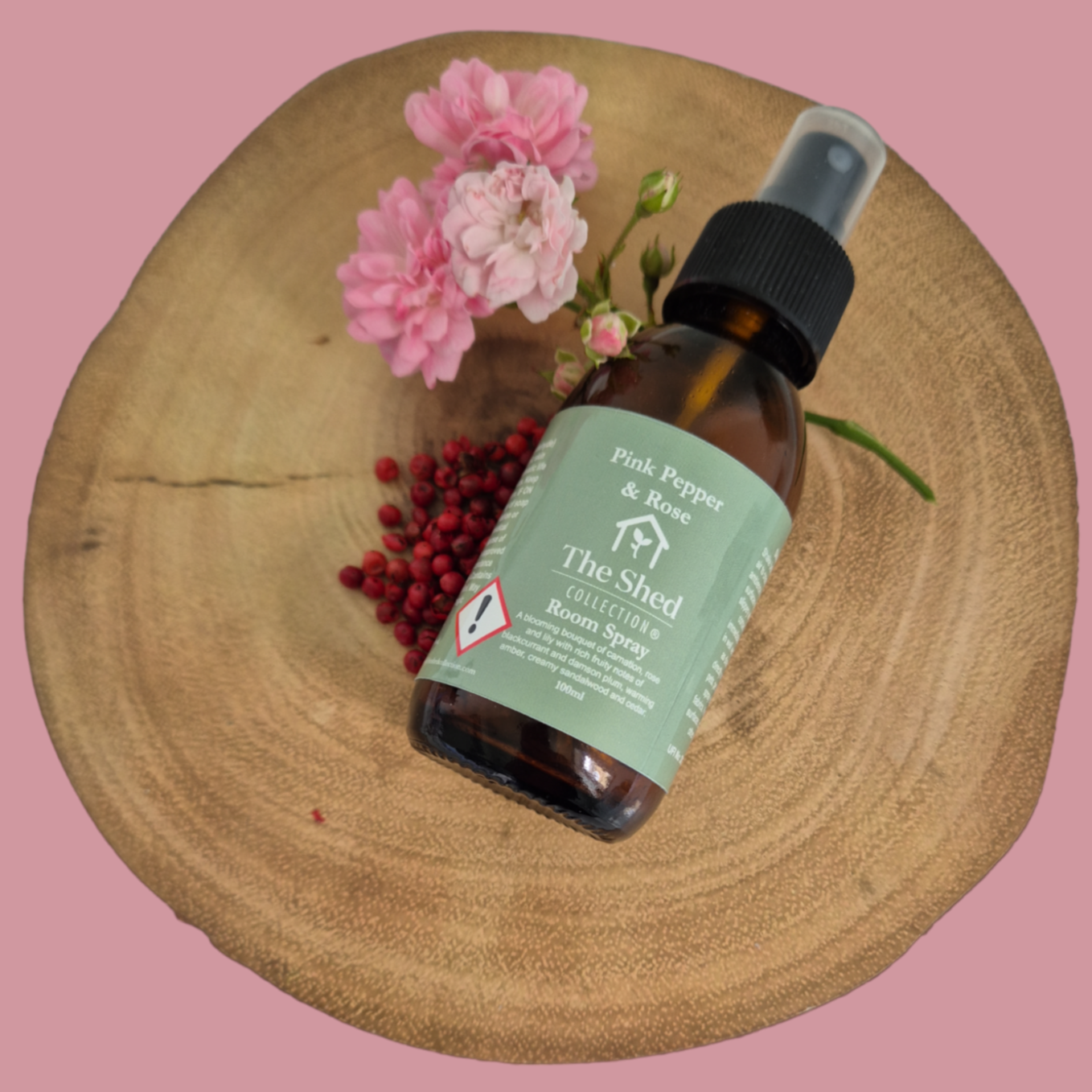 Picture shows a vegan friendly Pink Pepper & Rose room spray from The Shed Collection in a brown glass bottle with black spray lid on a log slice beside freshly cut miniature pink roses and some vibrant pink peppercorns.