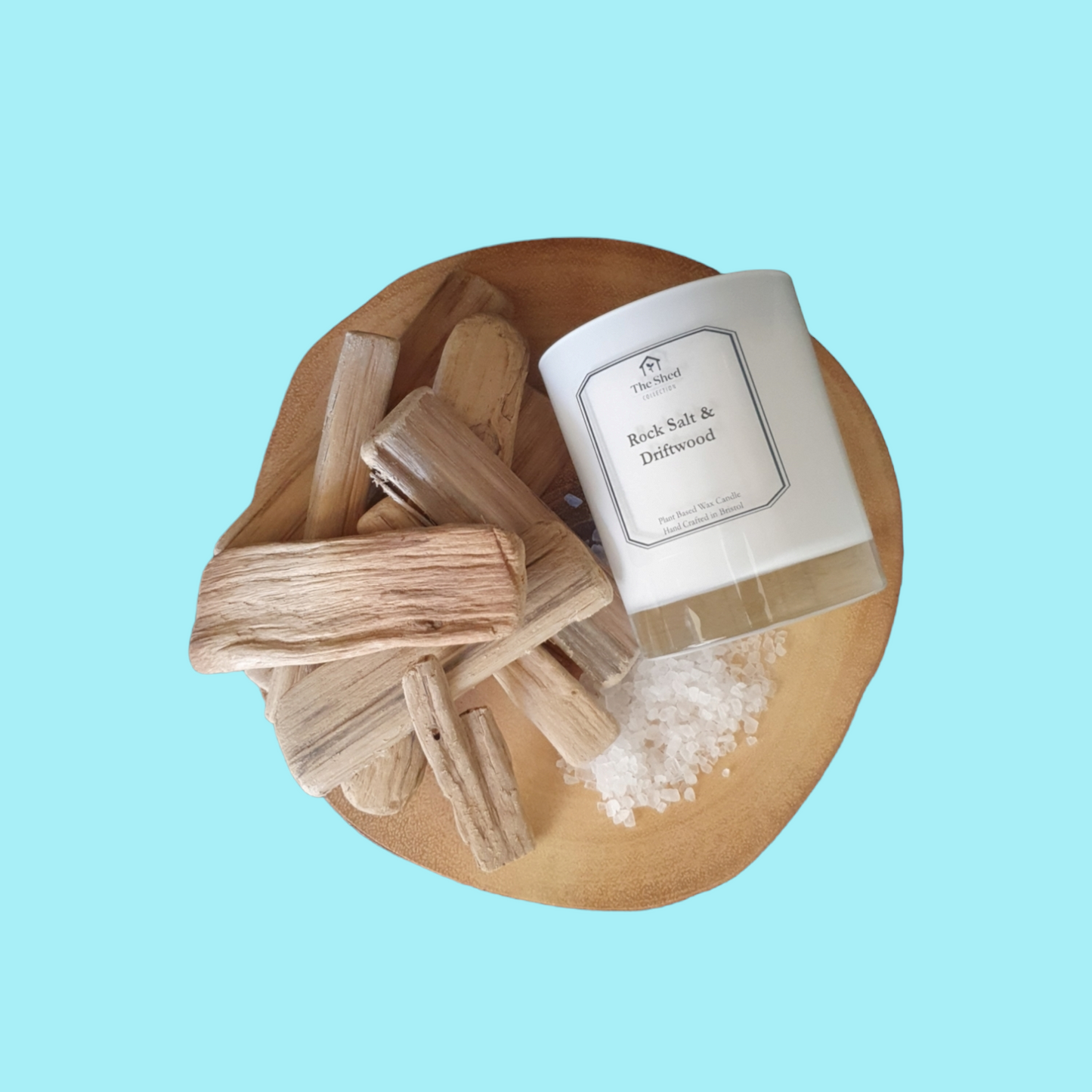 Picture shows a vegan friendly Rock Salt & Driftwood candle from The Shed Collection in a white container on a log slice beside pieces of driftwood and sea salt granules.