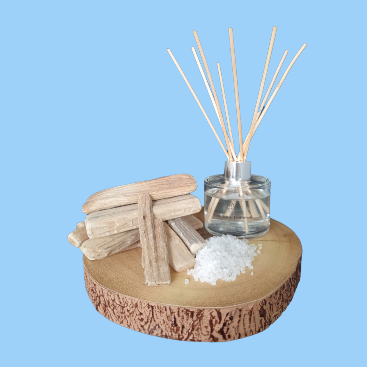 Picture shows a vegan friendly Rock Salt & Driftwood Reed Diffuser from The Shed Collection on a log slice beside pieces of driftwood and sea salt granules.