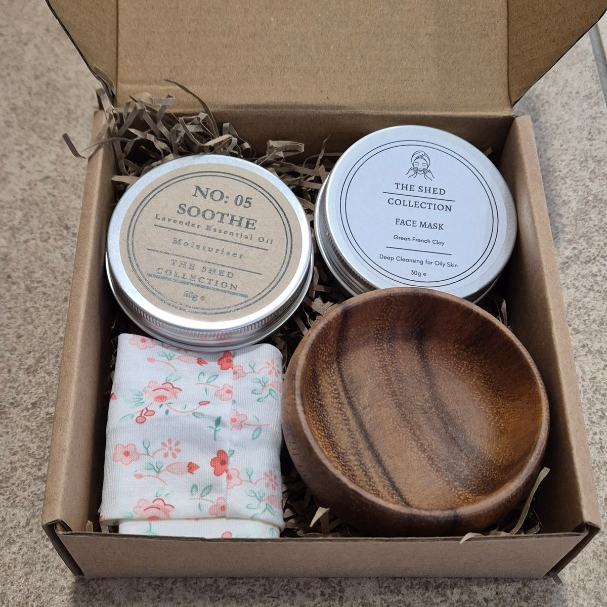 An open brown cardboard gift box filled with shredded paper and skincare items from The Shed Collection, including a wooden bowl, a floral headband, and two round metal tins labelled Soothe and Face Mask.