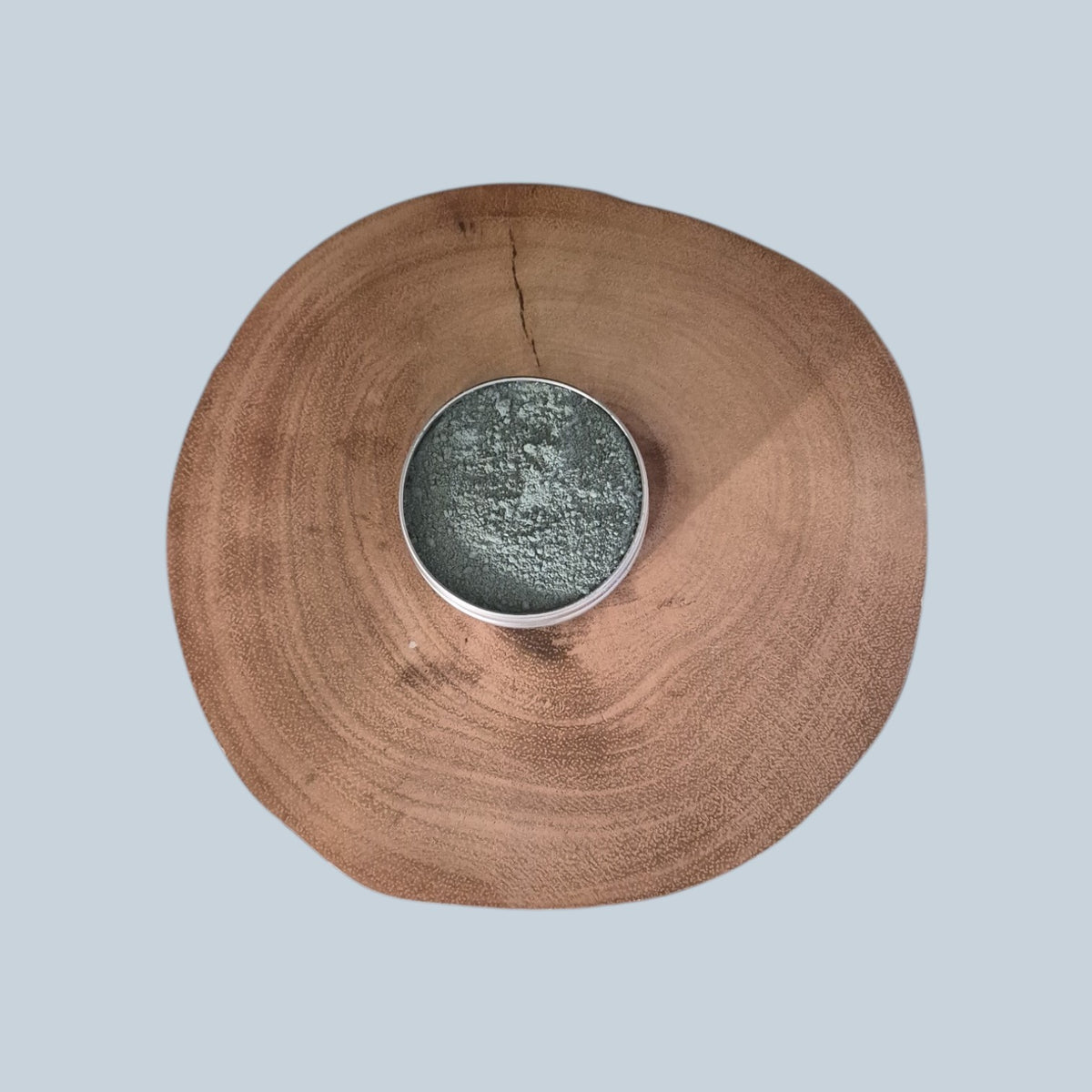 A round silver tin filled with textured gray-blue clay face mask sits centered on a circular natural wood slice against a solid light blue-gray background.