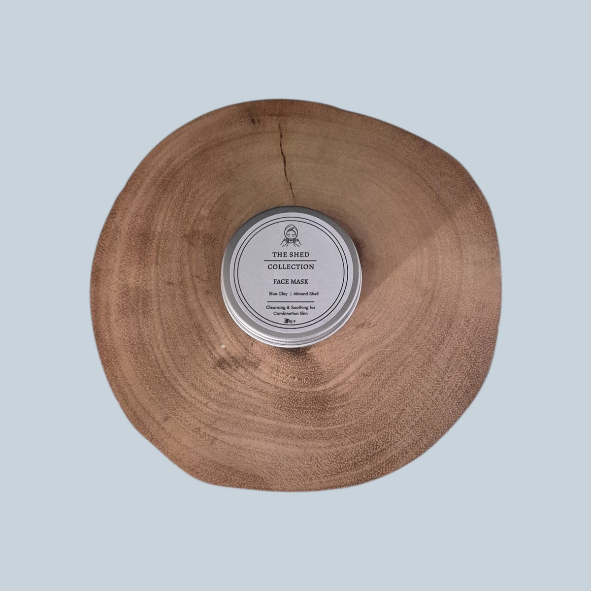A round silver tin of The Shed Collection Blue Clay & Almond Shell Face Mask tin placed in the center of a rustic, circular natural wood slice, set against a solid light blue-gray background.