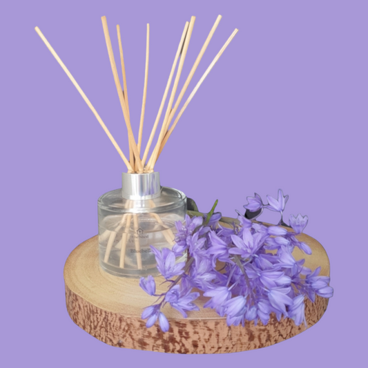 Picture shows The Shed Collection's vegan friendly Bluebell reed diffuser in a clear glass bottle with silver lid  
7 reeds sitting on a log slice surrounded by a bunch of Bluebells freshly picked from their Bristol garden