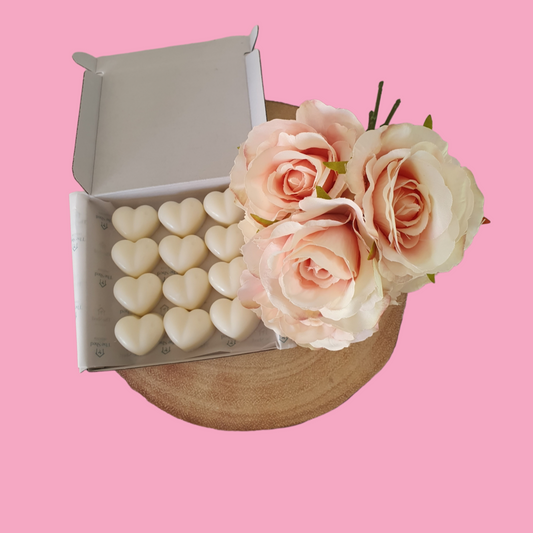 12 heart shaped wax melts in a vellum lined white box beside a bunch of pink roses. 