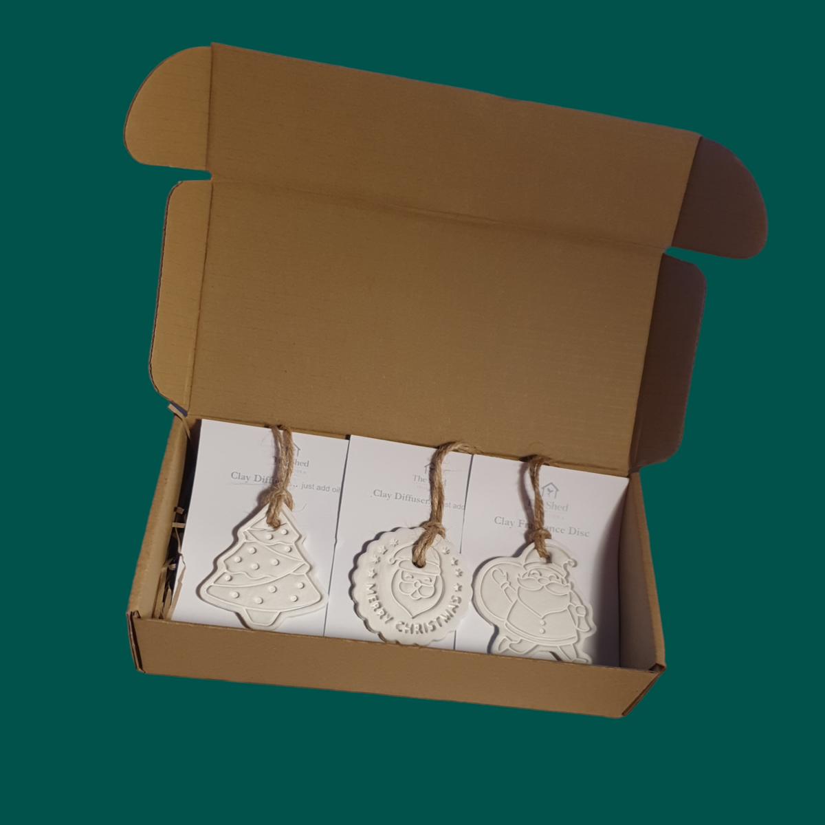 An open box of 3 clay shapes with brown natural cord hangers attached to white card. Shapes are Christmas Tree, Santa with Sack & round disc with a Santa face and the words Merry Christmas