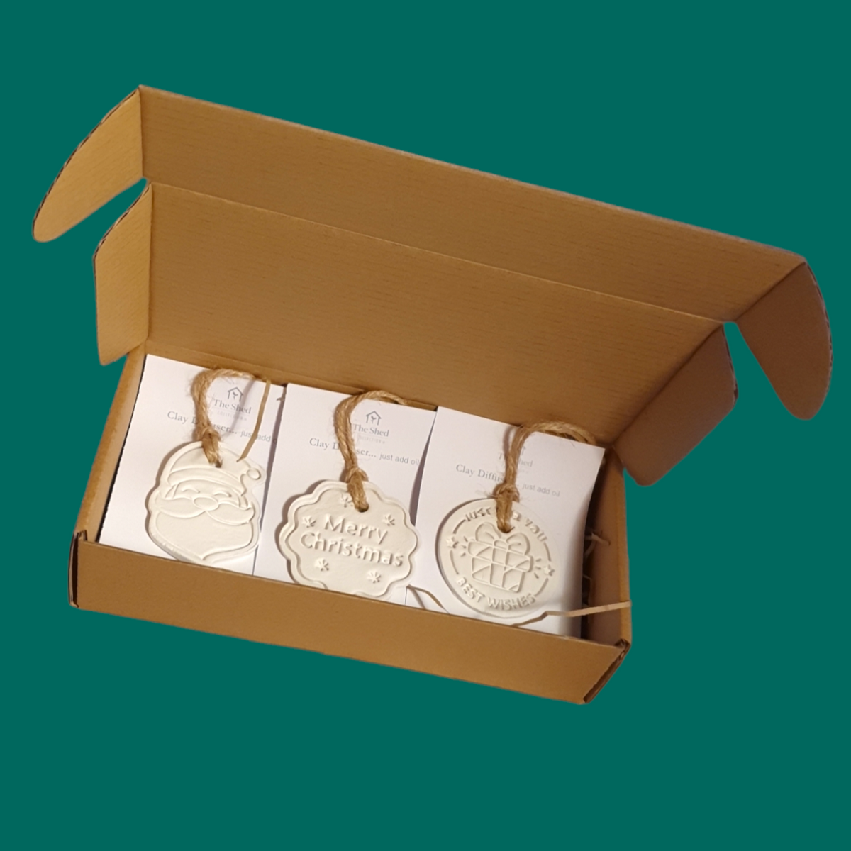 An open box of 3 clay shapes with brown natural cord hangers attached to white card. Shapes are a Santa Face, crinkled edged disc with the words Merry Christmas & round disc with a wrapped present & the words Just for You Merry Christmas