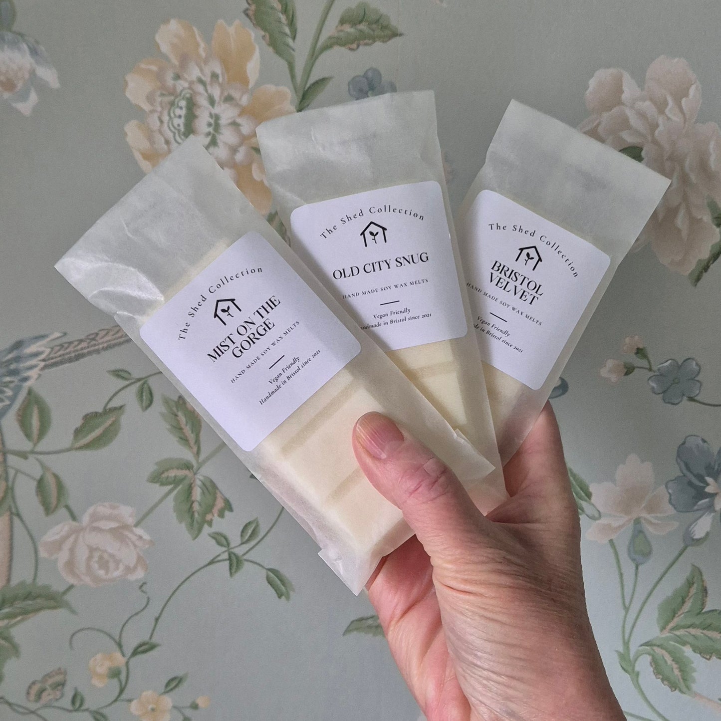 Hand holding three wax bars from 'The Shed Collection' against a floral-patterned background