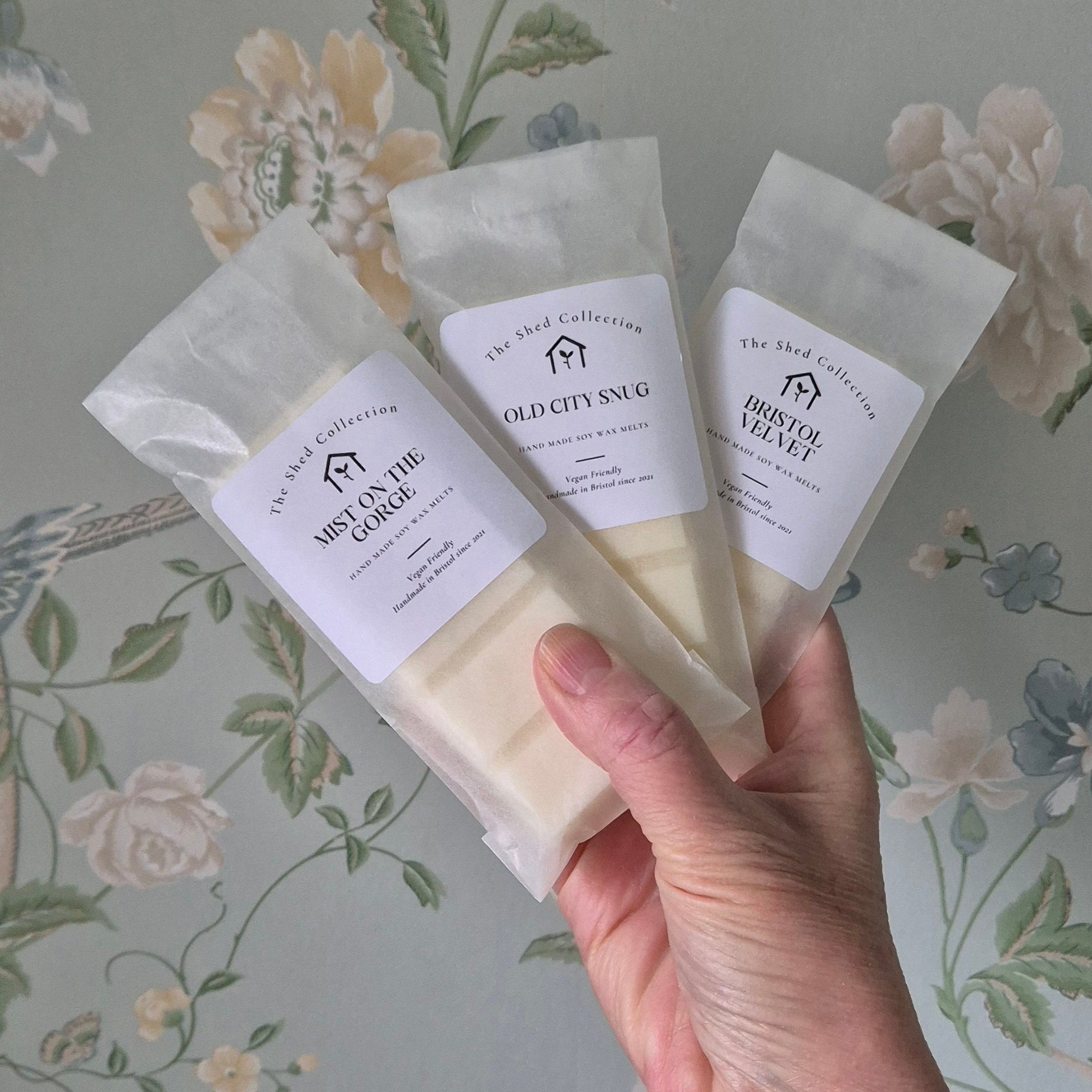 Hand holding three wax bars from 'The Shed Collection' against a floral-patterned background