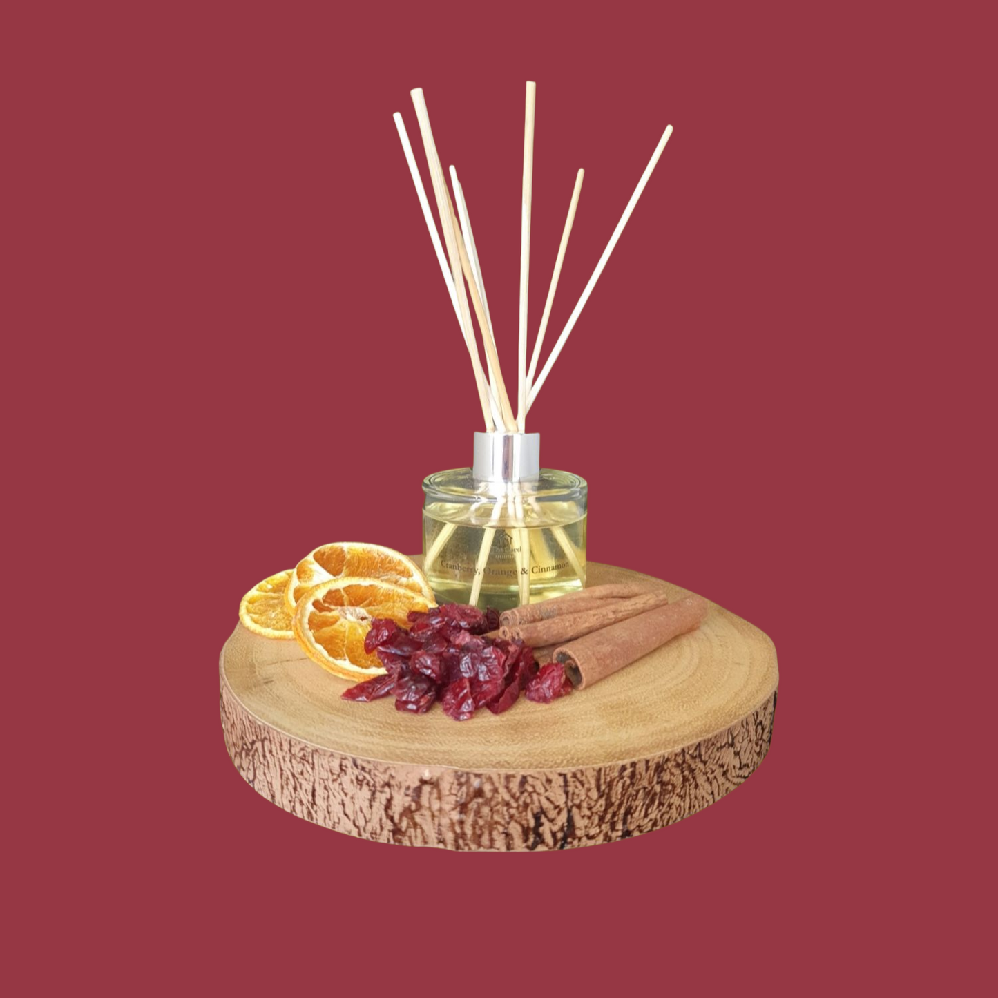 Reed diffuser with cinnamon, orange slices, and dried cranberries on a wooden coaster against a red background