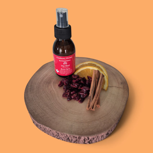 A small amber spray bottle with a black sprayer and a pink label reading Cranberry Orange & Cinnamon, styled on a round wooden slice with dried cranberries, cinnamon sticks, and a orange slice against a solid orange background.