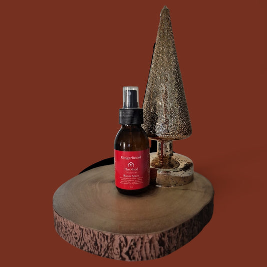 An amber glass spray bottle with a red label reading GINGERBBREAD The Shed Room Spray, styled on a rustic wood slice next to a decorative metallic Christmas tree against a solid rust-brown background.