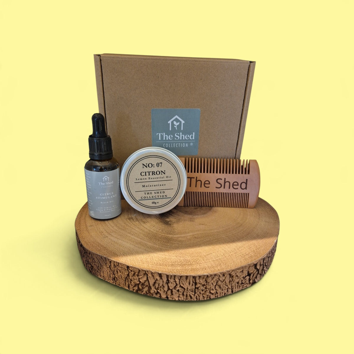 A curated beard care gift set from The Shed Collection, in a brown cardboard gift box, a small dropper bottle of beard oil, a tin of moisturiser, and a comb, all arranged on a rustic wood slice against a pale yellow background.