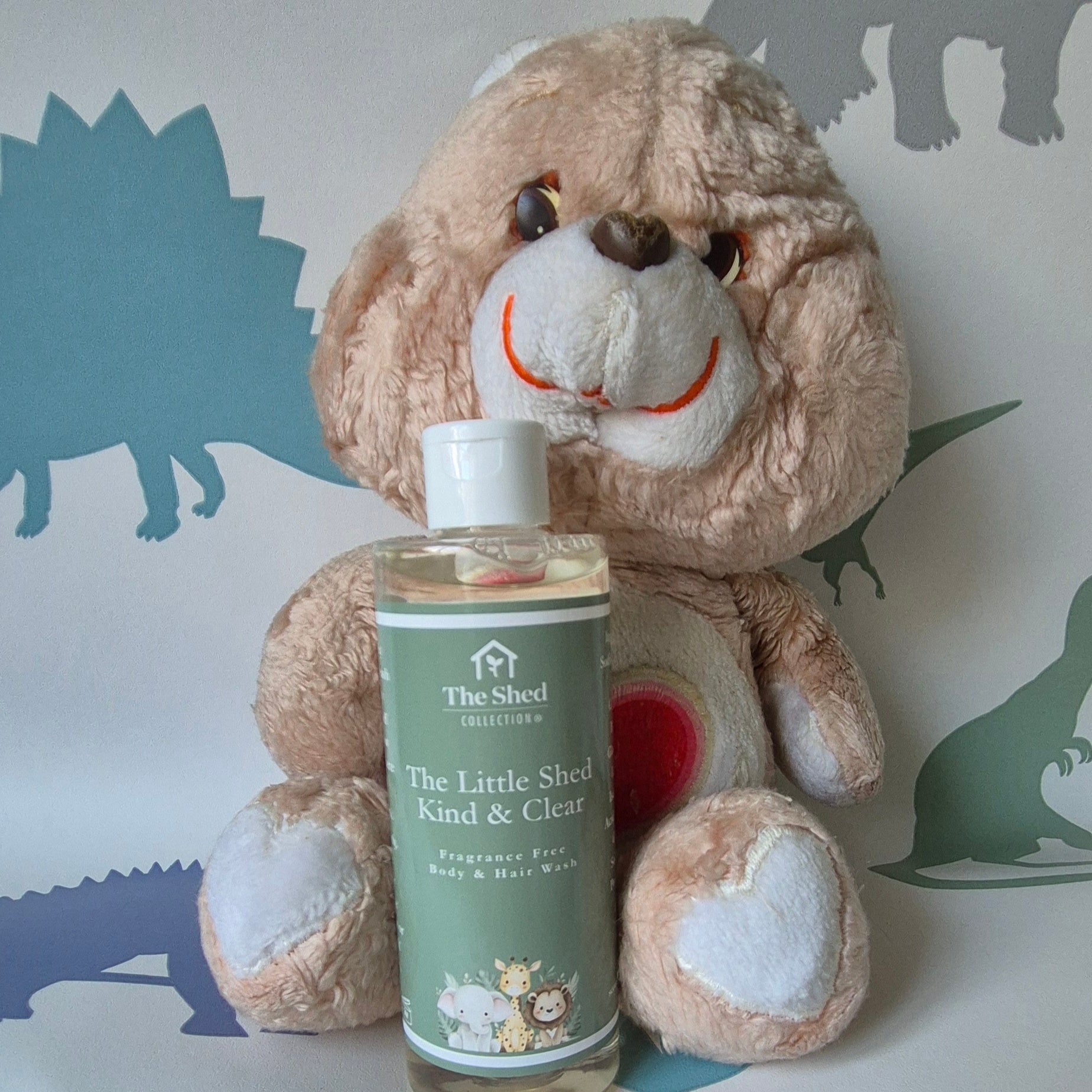 A bottle of The Shed Collection 'The Little Shed' Kind & Clear Fragrance Free Body  & Hair Wash, placed in front of a beige plush teddy bear against a white background with blue dinosaur silhouettes.