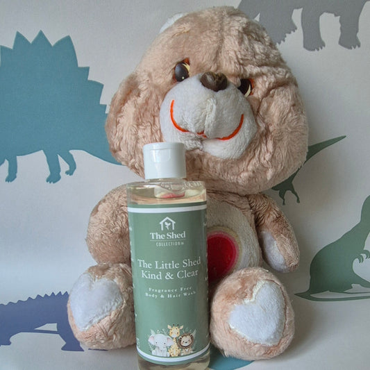 A bottle of The Shed Collection 'The Little Shed' Kind & Clear Fragrance Free Body  & Hair Wash, placed in front of a beige plush teddy bear against a white background with blue dinosaur silhouettes.
