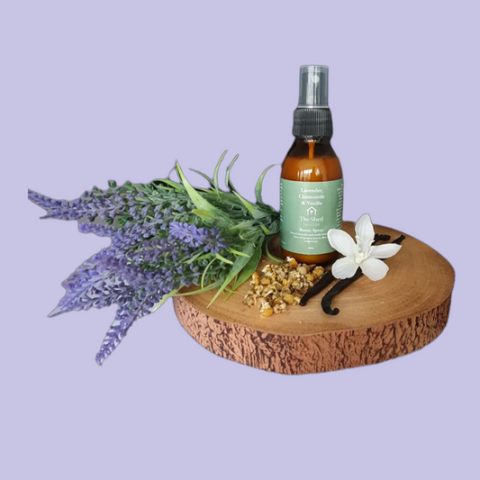 Picture shows a vegan friendly Lavender, Chamomile & Vanilla room spray from The Shed Collection in a brown glass bottle with black spray lid on a log slice with a bunch of lavender, two vanilla pods and some dried chamomile leaves.