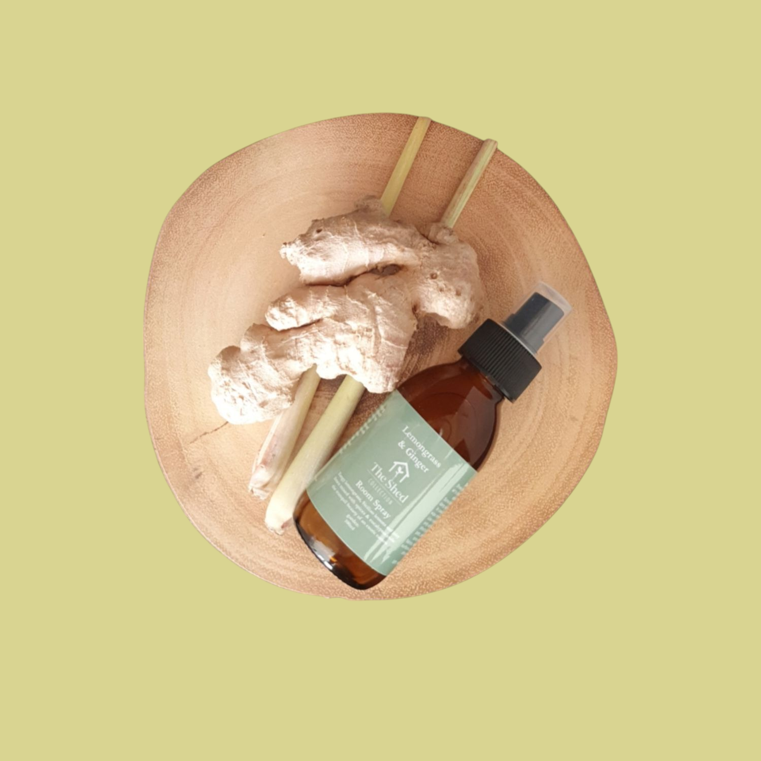 Picture shows a vegan friendly Lemongrass & Ginger room spray from The Shed Collection in a brown glass bottle with black spray lid on a log slice beside pieces of fresh lemongrass and ginger.