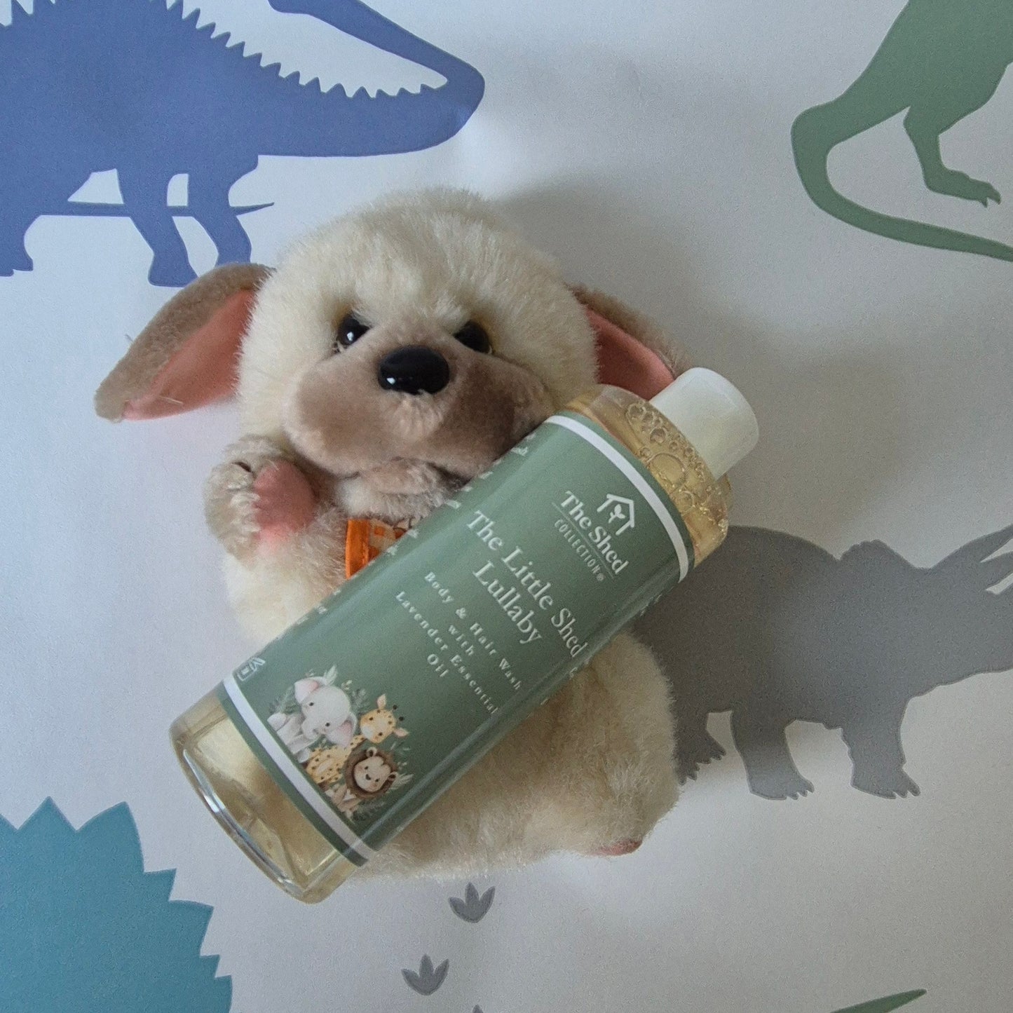 A bottle of 'The Little Shed' Lullaby hair & body wash placed on top of a small plush puppy toy, set against a white background with dinosaur silhouettes.