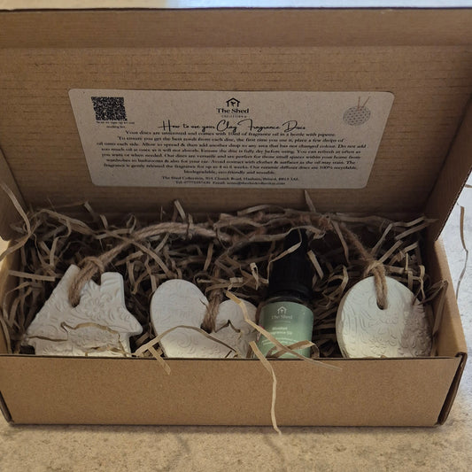 An open brown cardboard gift box filled with shredded paper and containing a small green bottle and three white ceramic ornaments, with a printed instruction card inside the lid.