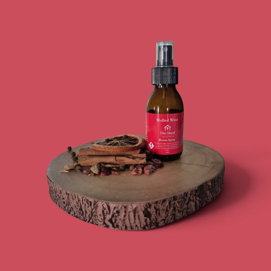 An amber glass spray bottle labeled Mulled Wine Room Spray stands on a round wood slice with dried botanicals, against a solid pink background.