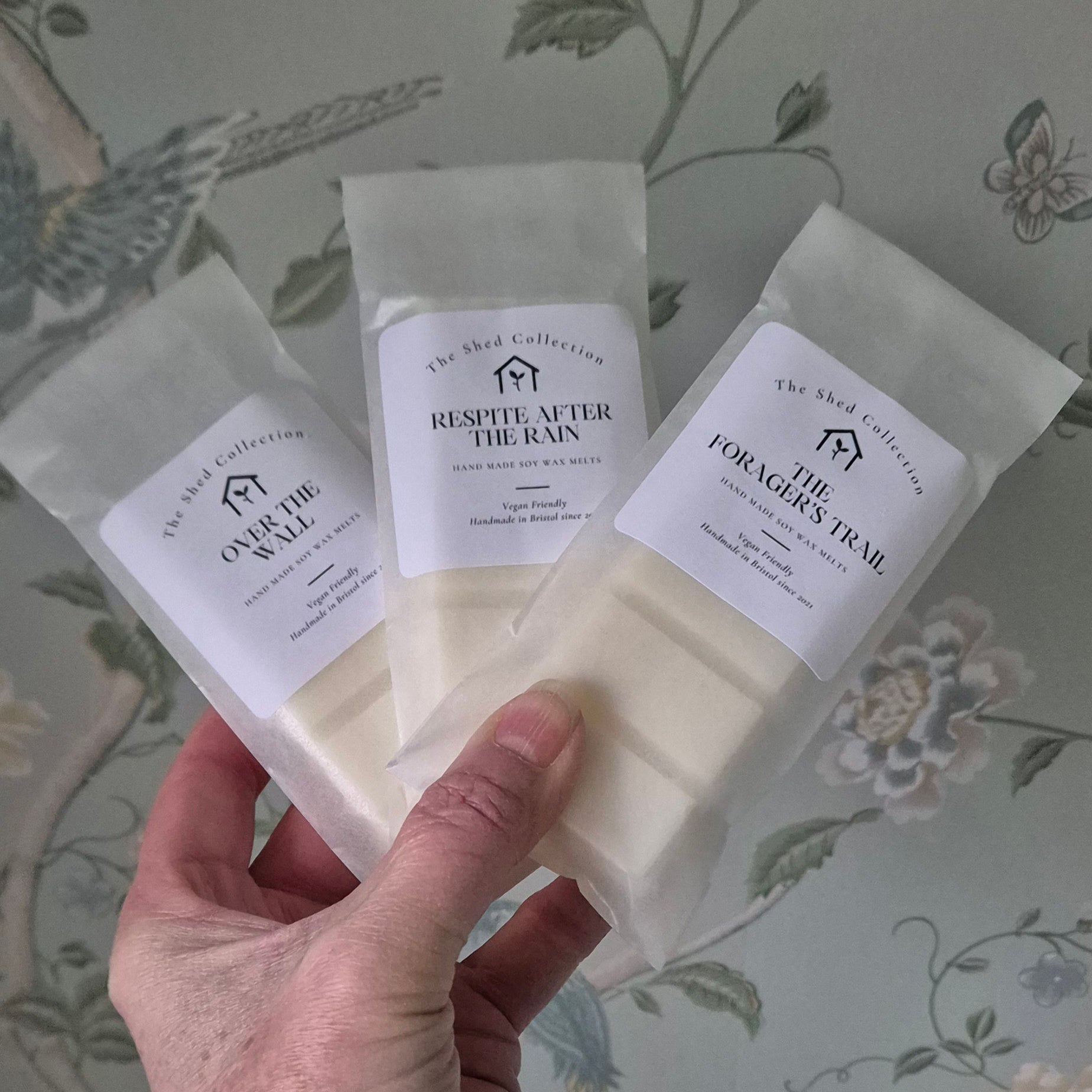 Hand holding three wax melt snap bars with labels against a floral-patterned background. Labels are Over the Wall, Respite After the Rain & The Forager's Trail