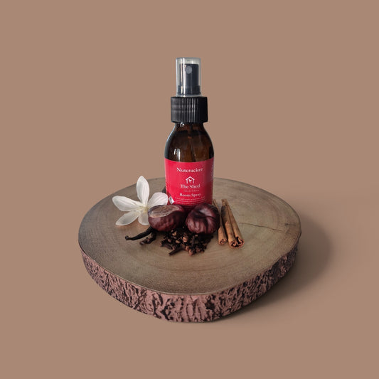 A small amber spray bottle with a black pump and a red label reading Nutcracker Room Spray sits on a round wood slice. The bottle is styled with a white flower, cloves, cinnamon sticks, and whole spices against a solid warm taupe background.