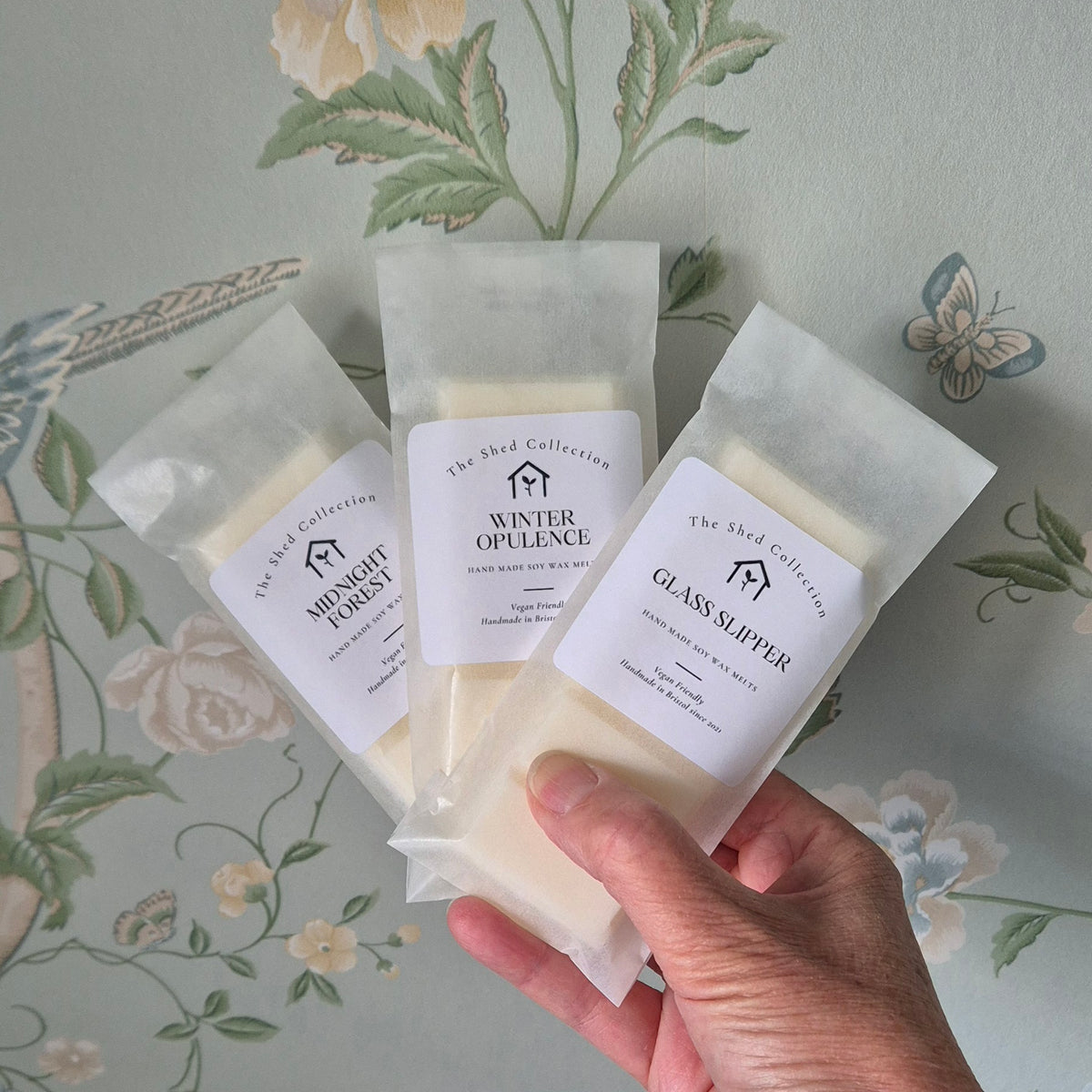 Three packaged bath salts from The Shed Collection are held in a hand against a floral wallpaper background. The packages contain off-white salts and are labeled 'Midnight Forest,' 'Winter Opulence,' and 'Glass Slipper.