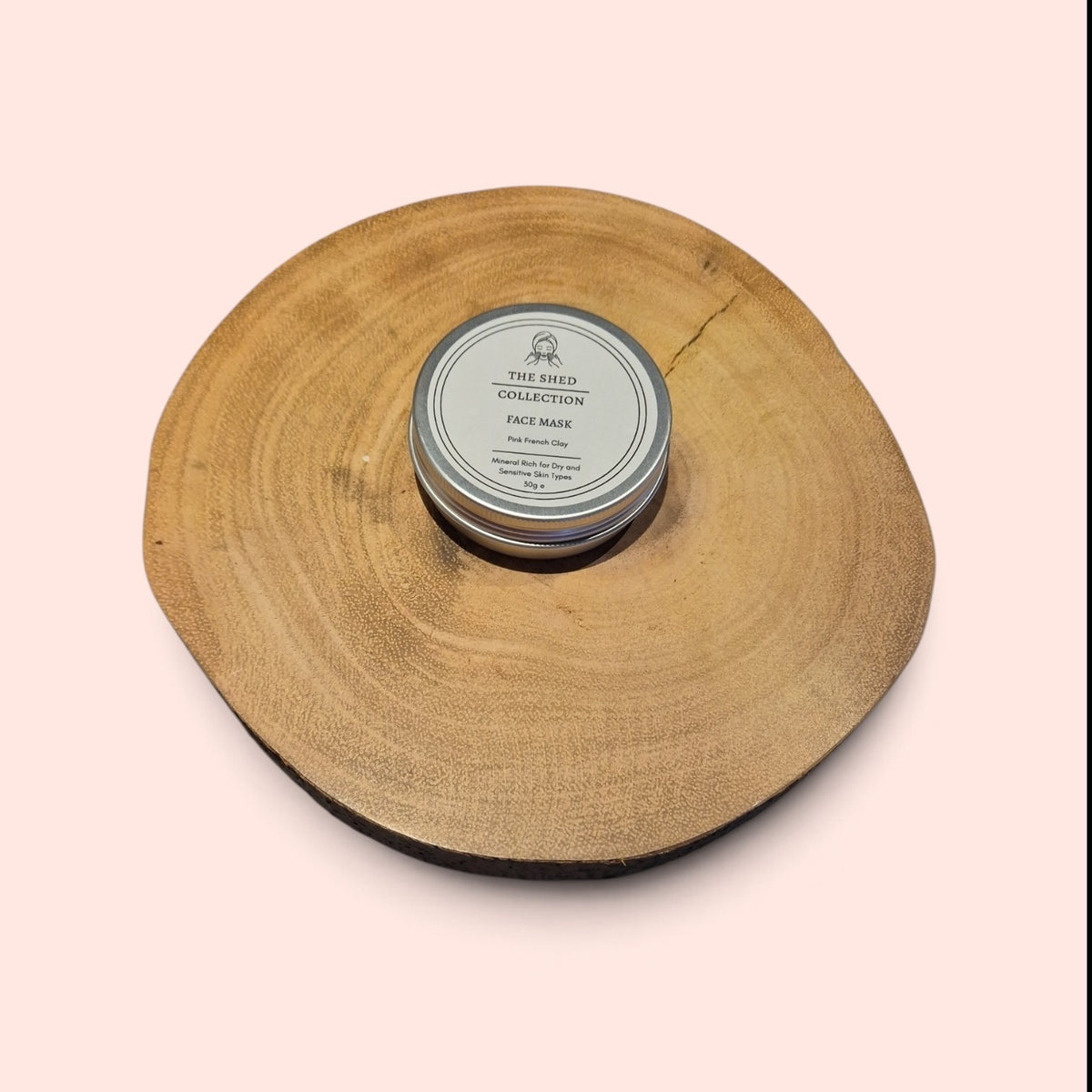 A round silver tin of The Shed Collection Face Mask placed in the centr of a natural wood slice coaster on a solid peale pink background.