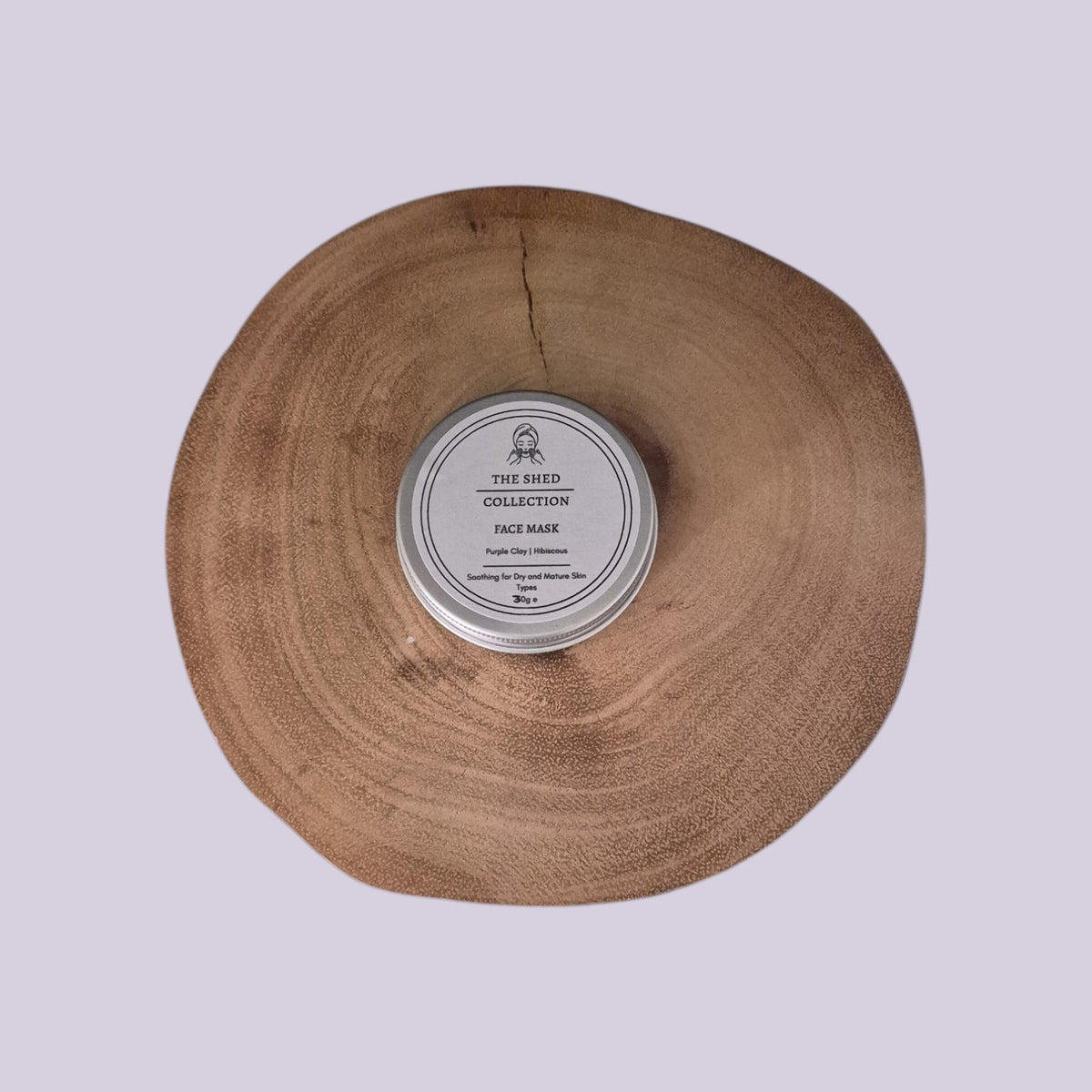 A round silver tin of The Shed Collection Face Mask Purple Clay & Hibiscus Face Mask placed in the centre of a large, rustic natural wood slice, set against a solid light lavender background.