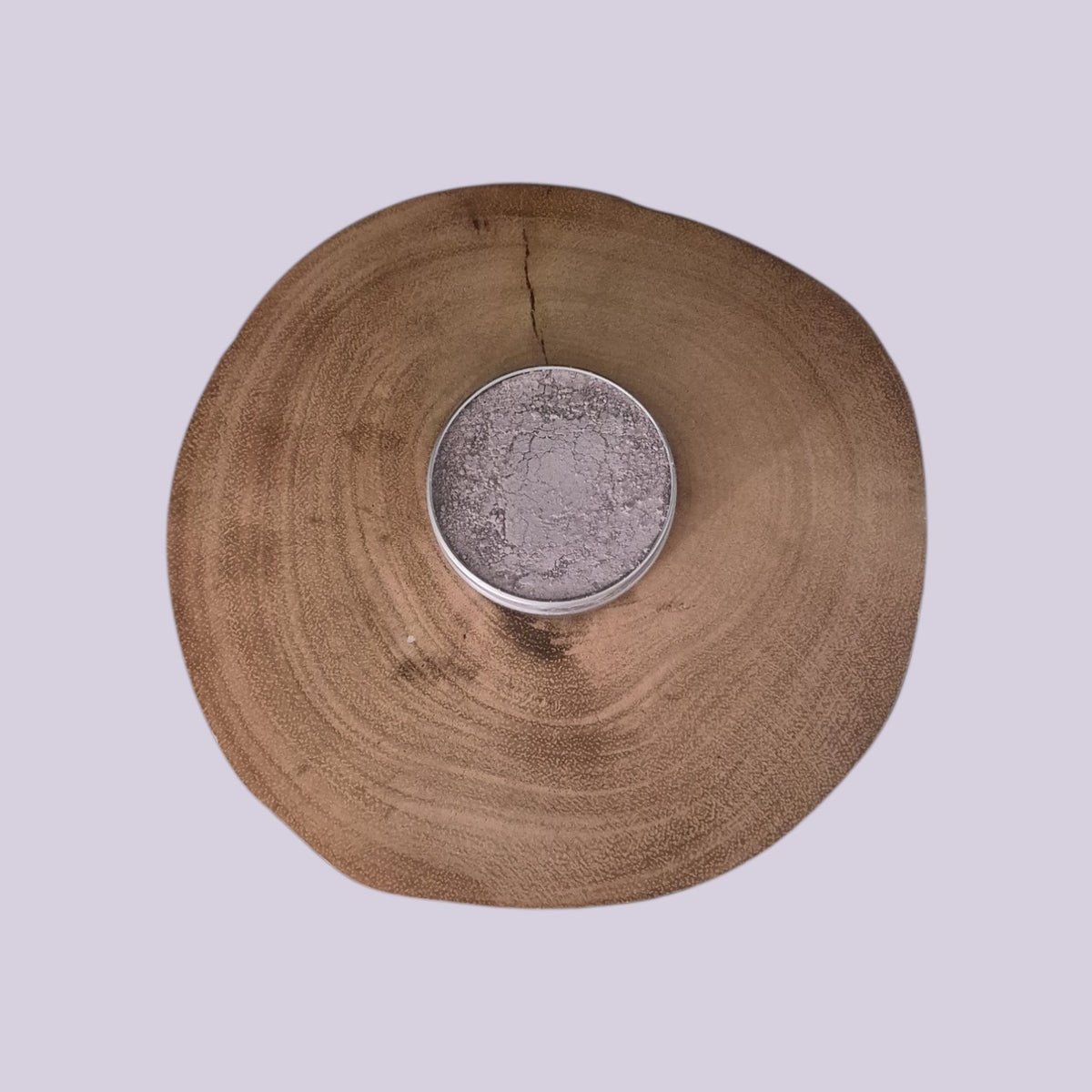A round silver tin filled with a light purple dry clay mask, placed in the center of a large natural wood slice on a pale lavender background.