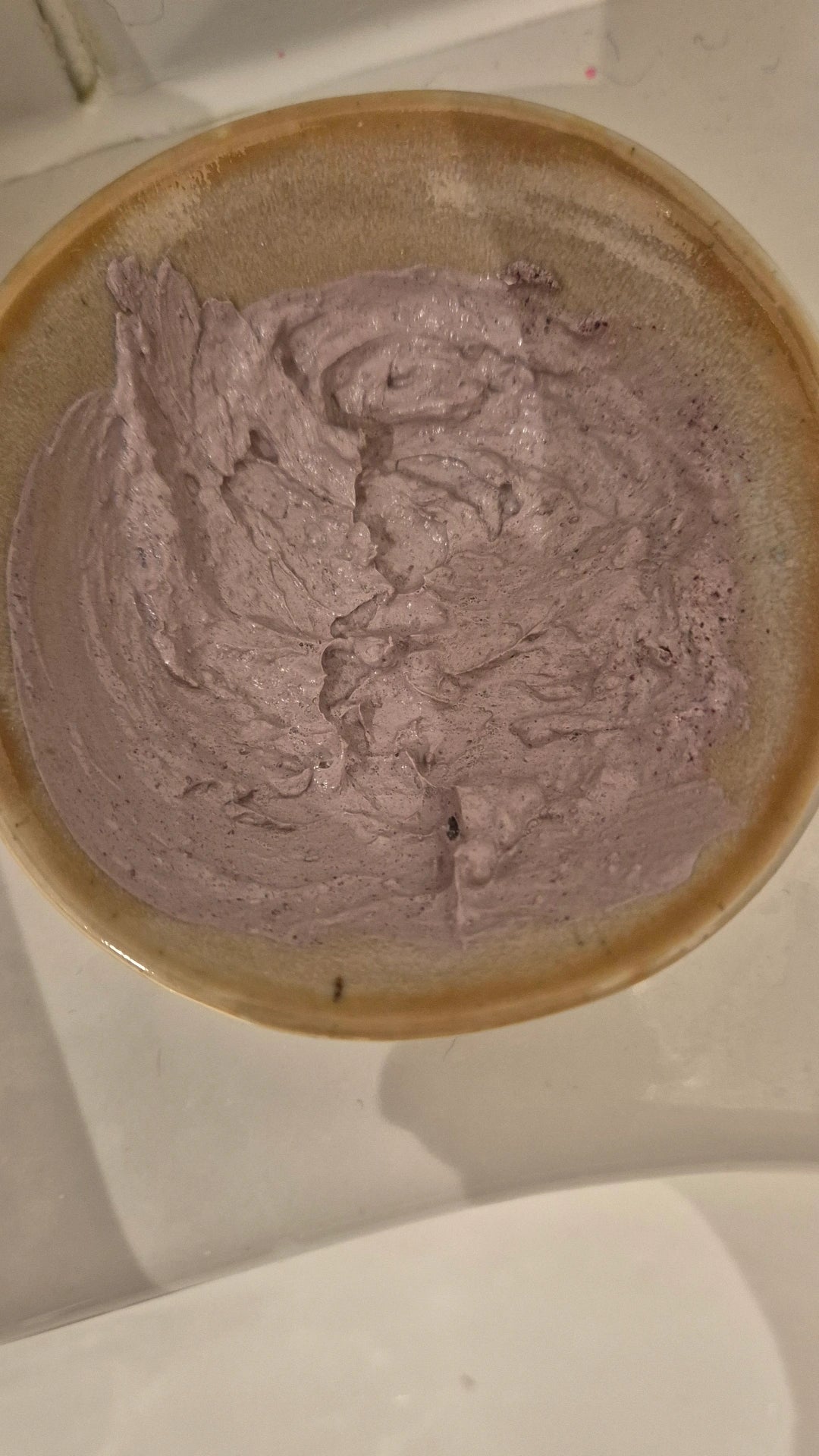 A close-up of Purple Clay & Hibiscus face mask mixed to a creamy lavender-purple swirled inside a rustic brown ceramic bowl on a white surface.