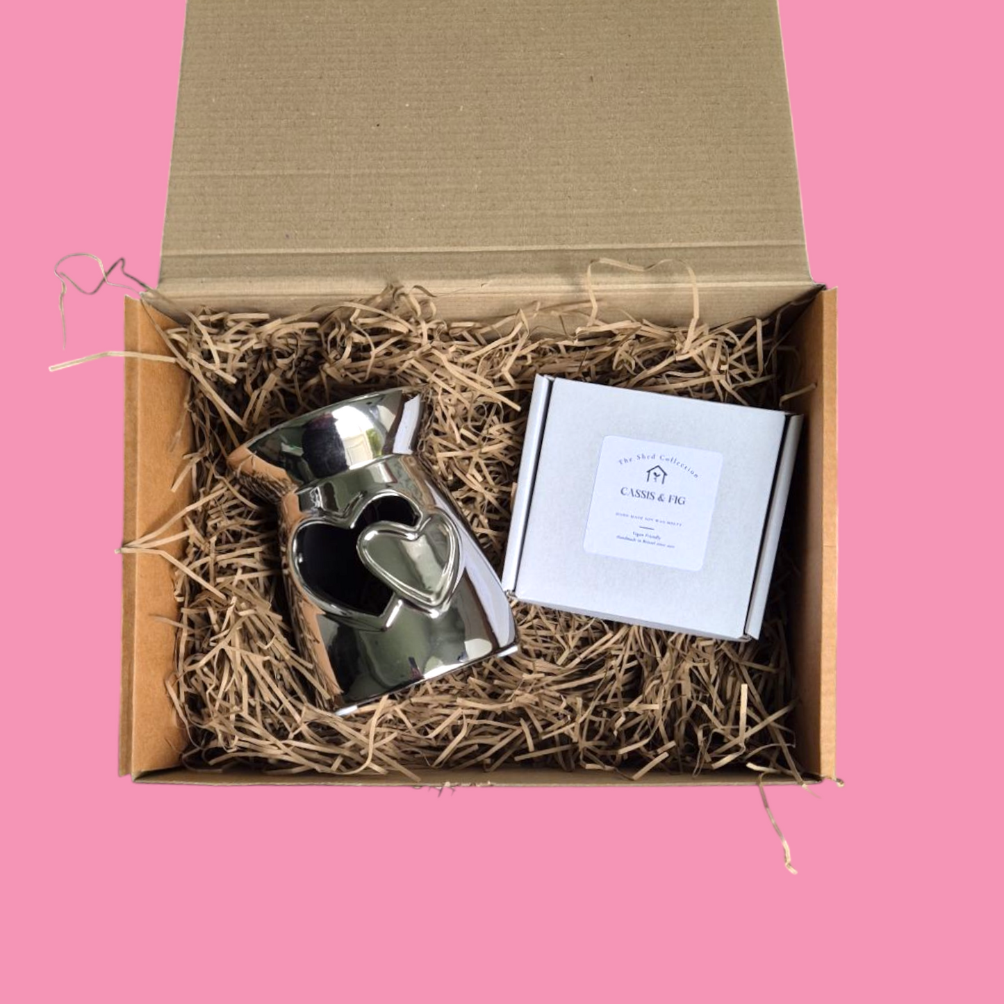 A silver heart-shaped wax melt burner is displayed in an open box with straw bedding, alongside a small white box with a label.