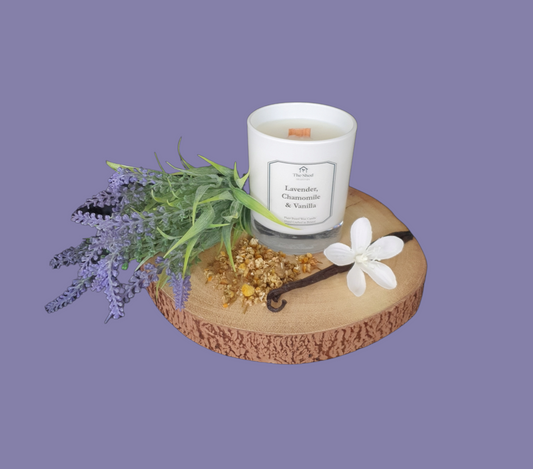 Picture shows a vegan friendly Lavender, Chamomile & Vanilla candle from The Shed Collection in a white container on a log slice with a bunch of lavender, two vanilla pods and some dried chamomile leaves.