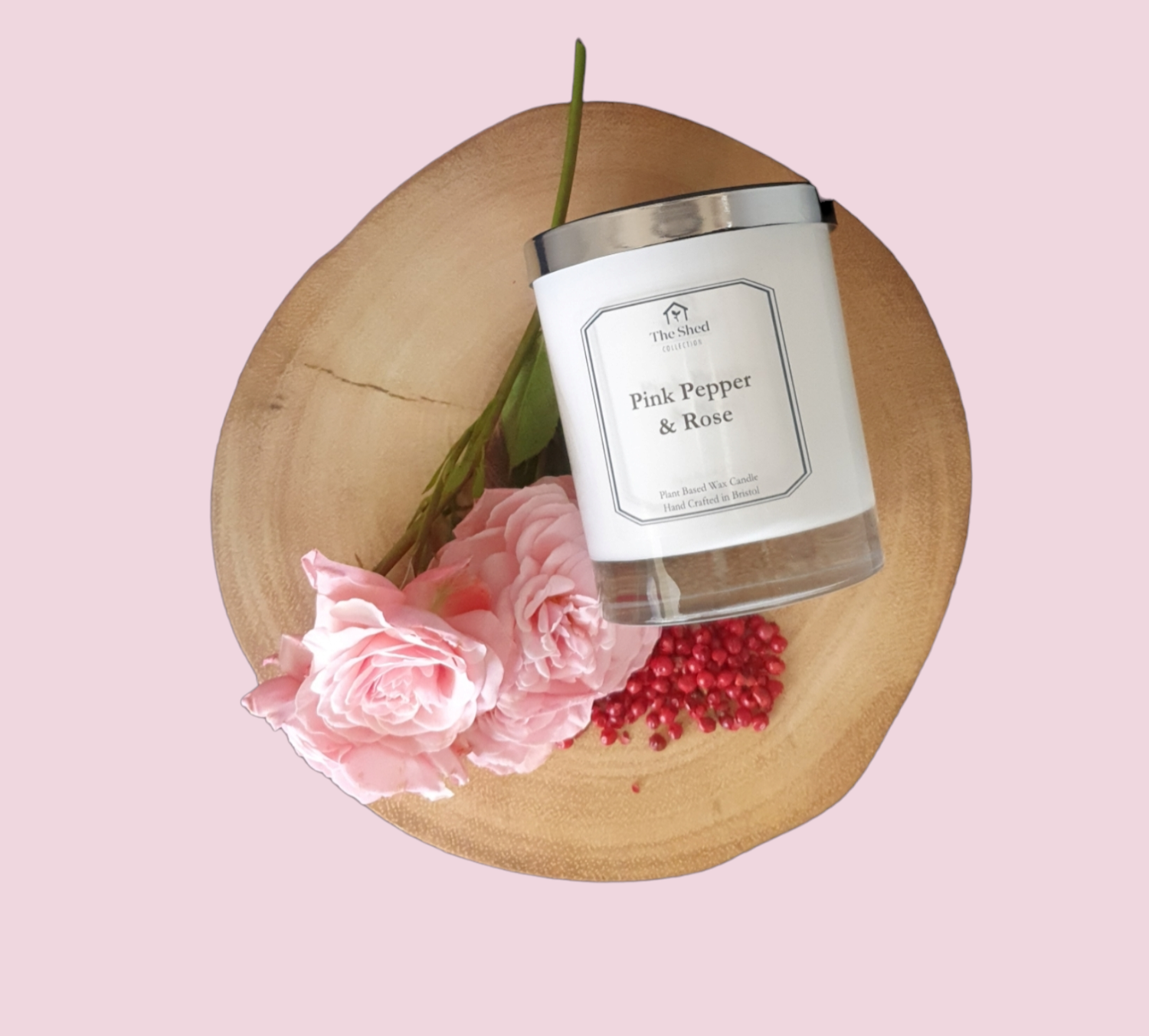 Picture shows a vegan friendly Pink Pepper & Rose candle from The Shed Collection in a white container with silver lid on a log slice beside freshly cut pink tea roses and some vibrant pink peppercorns. 
